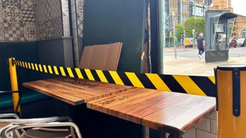 Inside a chipshop. Two tables with yellow and black tape in front. Window has been smashed away.