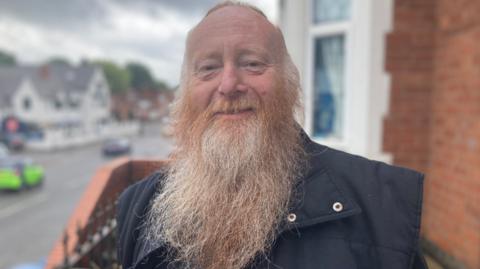 Resident Colin Millard, with a long beard, smiles at the camera outside a house 