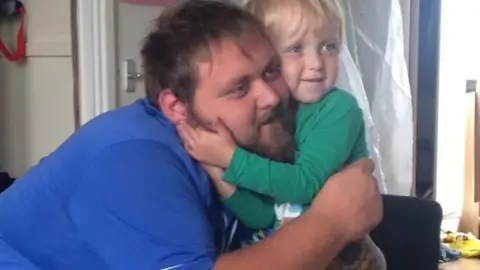 Supplied A young boy with blonde hair, wearing a green long sleeve top, cuddles a bearded man with brown hair, who is wearing a bright blue t-shirt.