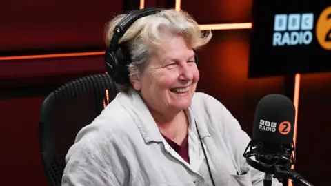 Sandi Toksvig is sitting an a BBC Radio 2 studio. In front of her is a black branded microphone and she is wearing headphones. The room is cast in dark orange lights. She is wearing a white shirt, red top and silver necklace. Toksvig  has short white and blonde hair and is smiling at someone off camera. 
