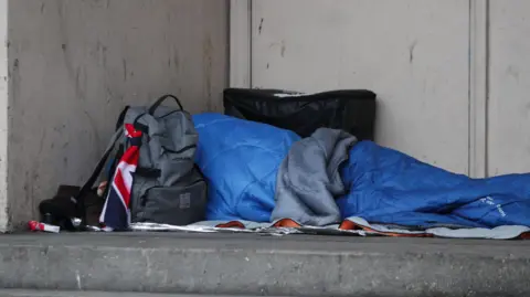 A picture of a person sleeping in a sleeping bag next to a doorway. There is a grey rucksack