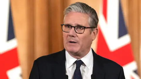PA Media Sir Keir Starmer has grey hair and black glasses and is wearing a white shirt, black tie and black suit jacket. Behind him is a wooden wall backdrop and two Union Jack flags