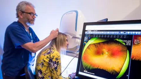 Sunderland University Woman with blonde hair and yellow top with blue flowers looking through eye machine. On the screen in front you can see a red and green scan of her eye. There is a man with glasses and a grey beard behind her wearing dark blue scrubs.