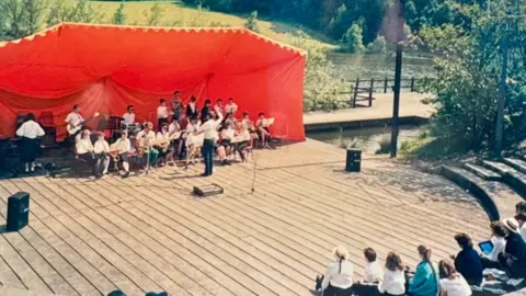 Matt Pearson A small crowd sits and watches a musical performance. They have a large red covering behind them as they play instruments on a wooden pontoon. Water and trees can be seen behind.