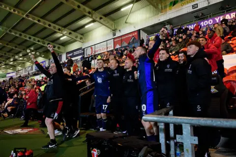 Chris Vaughan/Getty Images Football players and coaching staff stand along the touchline in a stadium, raising their arms in celebration. The group wears dark blue kits and black coats, with seated spectators and advertising boards behind them.