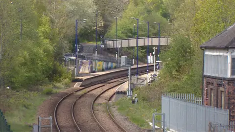 A view of the railway platform with some tracks leading up to it. There is a concrete bridge over the tracks and tall street lights lining either side. On the right there is a brick signal box. Trees surround the platform and tracks.