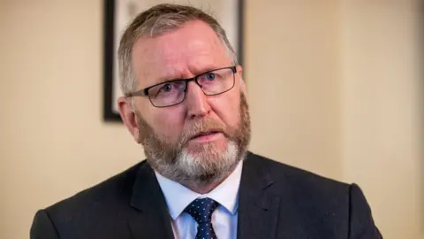 Doug Beattie, a man with greying fair hair and a matching beard, looks towards the camera with a serious expression. He is pictured in an office setting with pale yellow or cream walls. He is wearing black-framed glasses, a black suit jacket, a white shirt and a navy and white polka dot tie. 