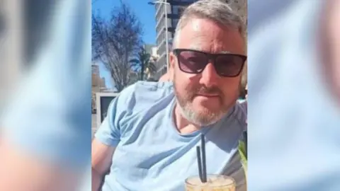Photo of David Rogers sat outside wearing sunglasses with a drink in front of him. He is wearing a blue t-shirt and has short grey hair and beard.