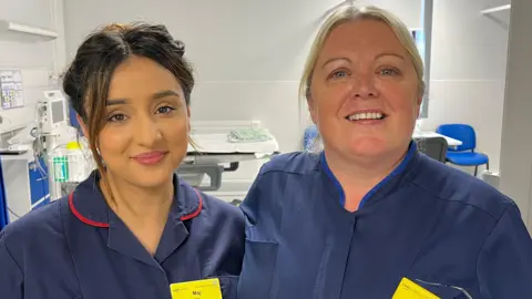 Maj and Clare standing with their arms around each others' shoulders and looking into the camera and smiling. They're both wearing dark navy blue nurses' uniforms - Maj, on the left, has red borders on the collar, and Clare, on the right, has a lighter blue collar. They're both wearing bright yellow NHS name badges - but you can't quite make out what's written on them. They're standing in the entrance to one of the bed bays - a hospital bed is just visible beyond them, and the room has bright white walls and floors. 