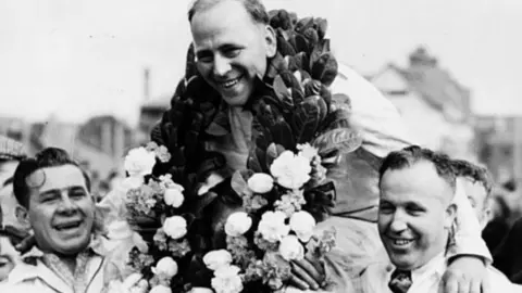 A black and white image of British racing driving Reg Parnell being carried by his all male Maserati pit crew, following his victory in Jersey, May 9th 1947. He is wreathed with flowers and smiling broadly. They are all wearing overalls. 