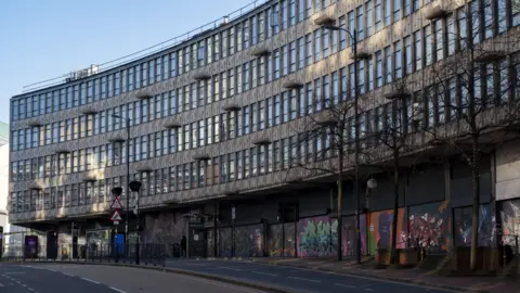 Mike Kemp/Getty Images A curved building with grey walls and rectangular windows. There is graffiti on the ground floor.