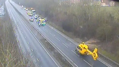 National Highways A highways camera image of the A14 near Bury St Edmunds. A yellow helicopter and emergency services vehicles can be seen on the road. 