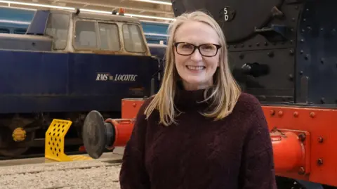 Sarah Price has long blonde hair and glasses and is smiling and wearing a long maroon jumper. She is standing in front of a black and red locomotive and a blue locomotive inside a museum.