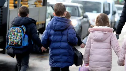 A group of children walking hand in hand down a a pavement next to a row of cars on the road to the left.