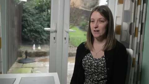 A woman in either her twenties or thirties with dark shoulder length hair. She is stood against the backdrop of some patio doors which lead out to a garden. She is wearing a black cardigan and flowery dress.