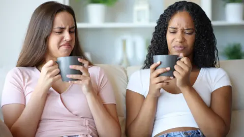 Getty Images Two women looking distastefully successful  to their mugs whilst sat connected  the sofa