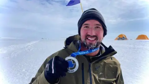 A man in Antarctica wearing a navy blue beanie, thick black gloves, and a green coat. He is smiling and holding a silver medal with a blue ribbon. Behind him is bright white snow, and yellow tents 