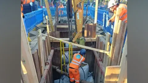 Thames Water Engineers carrying out repair work in a deep excavated trench around a large water main. A worker in high-visibility orange protective clothing stands at the bottom of the shored excavation beside exposed pipework and repair materials, while another worker operates machinery from the surface.