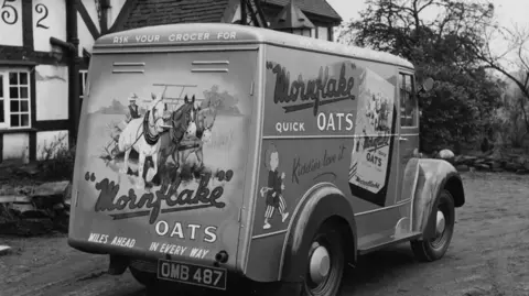 Mornflake A black and white image showing an old-fashioned van with Mornflake branding on the side. It is parked outside a timber-framed house.