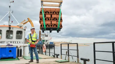 PA Media A man wearing jeans, white hard hat and a high vis fluorescent jacket is standing on a boat. He is operating a crane which has a pallet of clay bricks on it. 