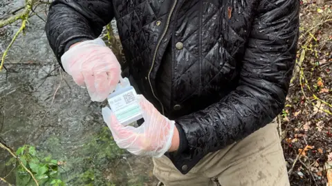 Kate Bradbrook/BBC A person wearing a black coat and beige trousers is stood in the middle of the image holding a piece equipment used to test water. They are wearing gloves and holding onto the cleared bottle which has a label on the front with a QR code.