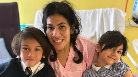Raina Heverin A woman sits in a hospital bed with two small children. One child is under each of her arms. 