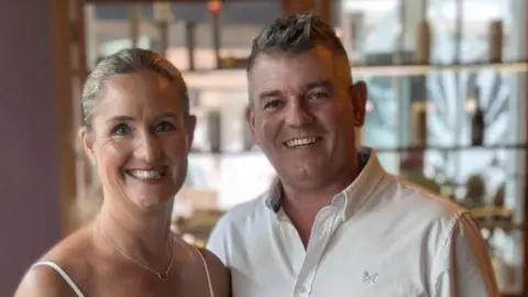 Supplied A man and woman stand side by side, smiling. The man wears a white shirt and has short grey hair, and the woman wears a white dress with thin straps. She has blonde hair tied back in a ponytail.