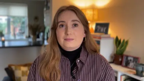 Nathan Turvey/BBC A picture of Emily Lynn, who has ginger hair and is wearing a striped shirt. There are pictures, a lamp shade and her kitchen in the background.
