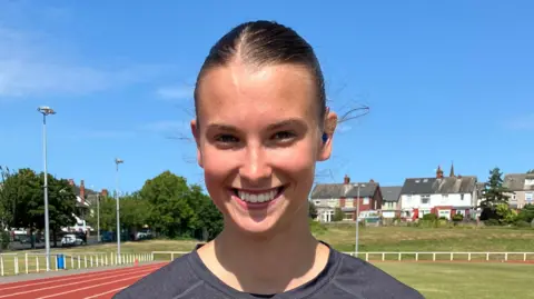 Jennie Dennett/BBC Honor smiling into the camera. She is standing in the same position as the previous image. The sky above her is bright blue and cloudless.