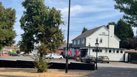 A brick pub painted white with several cars parked outside. benches with red umbrellas are outside the front of the pub.
A low brick wall seperates the car park from the pavement and road outside. The words The Craftsman are written on the side of the pub.