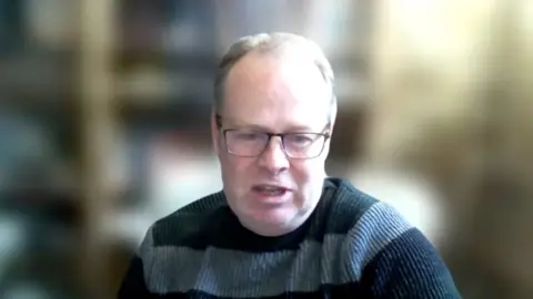 A man wearing glasses and a stripey jumper sits infront of a bookcase