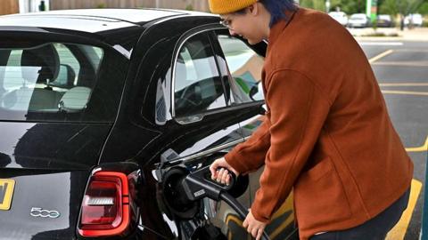 A woman wearing an orange coloured coat fills up a black Fiat 500 car with diesel at a petrol station