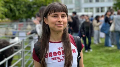 A woman with long brown hair and a fringe is wearing a red and white t-shirt and wearing a backpack. She is standing by a metal fence where animals are inside. 