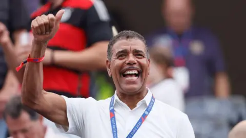 Chris Kamara waves from the stand before the UEFA EURO 2024 group stage match. He has short black hair and a wide smile.