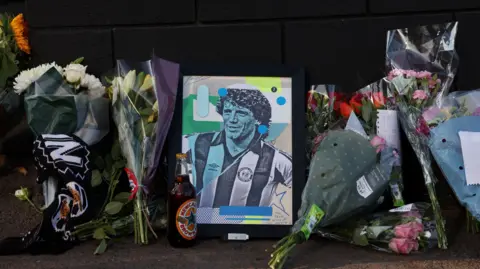 Bouquets of flowers leaning against a dark wall with a colourful print of Kevin Keegan when he was younger in a Newcastle shirt. There is a bottle of Newcastle Brown Ale next to it.