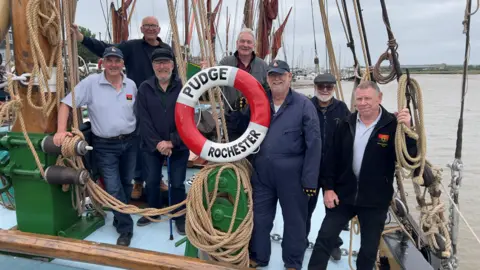 John Fairhall/BBC Seven men stand on the deck of Pudge. The various ropes, chains and the mast can be seen. They are wearing a mixture of darkly-coloured fleeces, jackets and polo shirts.