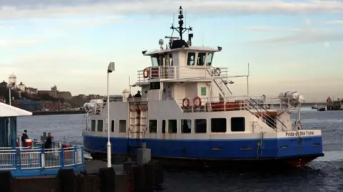 The Shields Ferry, a white and blue boat which has Pride of the Tyne written on the back of it. It has an indoor level, with an outdoor level above it. It is at one of the landings which is surrounded by a blue and white fence. Two people are waiting by the barriers. 
