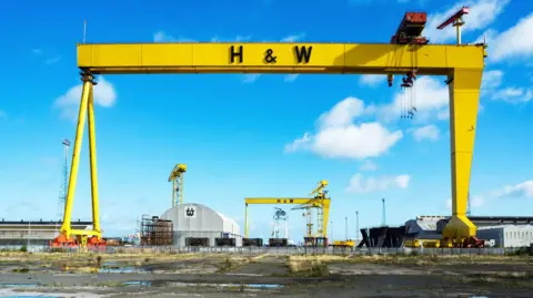 GETTY IMAGES Um dos guindastes da Harland and Wolff. É amarelo, com letras pretas “H & W”. O céu atrás do guindaste é azul. Vários edifícios podem ser vistos ao fundo.