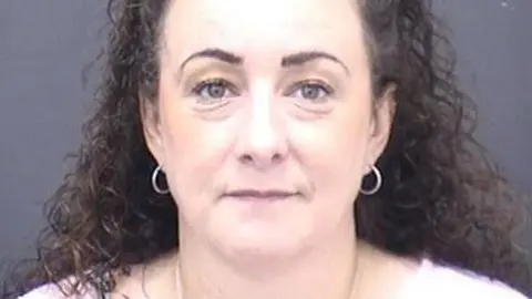 A custody photo of Emma Charlotte Amey, a woman with long dark curly hair and wearing small hoop earrings.