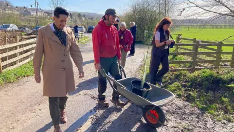 A man pushing a wheelbarrow with a black pot containing a tree sapling with two people walking either side of him.