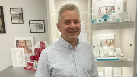 BBC jeweler Andrew Clayton stands inside his shop, in front of display cases of jewellery. He is smiling and has short gray hair and is wearing a white and blue striped shirt.