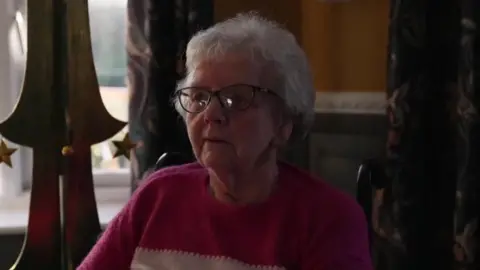 Shirley Medd sitting on a chair with large Christmas ornaments behind her. She has short, curly, grey hair and is wearing black-rimmed glasses and a pink and white sweater.