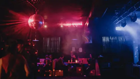 Ameila Read The inside of Lubber Fiend during a club night. The room is dark with red and blue lights shining inside. There is a disco ball to the left of the ceiling and people dancing underneath it. There are people sat around a table towards the back.