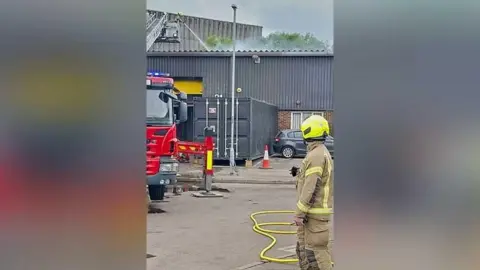 Smoke is seen rising from an industrial building as a firefighter in uniform watches on. There is a hose on the ground beside him.