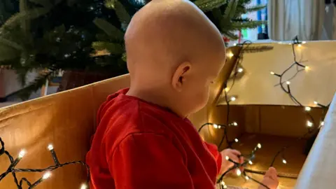 Maddie Biggs A young baby is wearing a red top and is sitting in a cardboard box while looking at sparkly Christmas lights