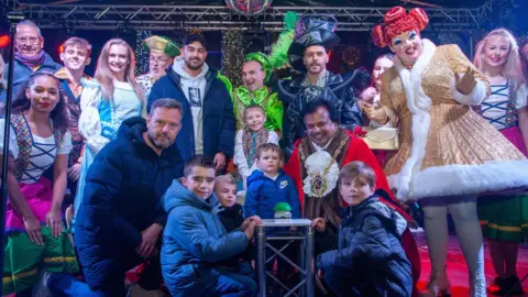 Lucy Taylor Newsquest A large group of people stand on a stage in the centre of Ipswich. Among them is Ipswich Town FC chief executive Mark Ashton, who is kneeling on the front row, and panto star Louie Spence, who is at the back dressed in a vivid green outfit for his part in Jack and the Beanstalk at the Wolsey Theatre. There are other pantomime characters on the stage along with local childen