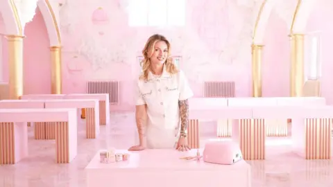 A large room painted completely pink with pink tables and arches with gold columns. A woman is stood in the middle of the room with nail products smiling at the camera