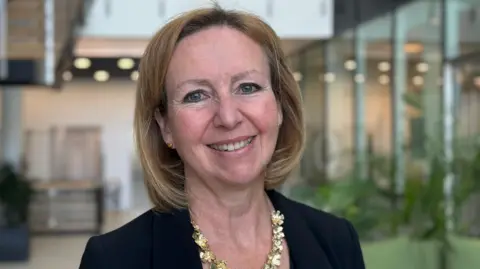 Jane Hutchins is standing in a room which has large glass windows and plants on the right. Hutchins has short light brown hair and is wearing a black blazer and gold necklace.