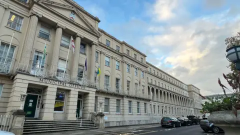 The front of Cheltenham Borough Council's Municipal Offices. It is a beige-grey four storey stone building and is very wide. There are car parking spaces in front of it and a cloudy blue daytime sky in the background. Part of a tall stone planter can be seen in the foreground of the image on the right. Four flags are flying on the front of the buiding.