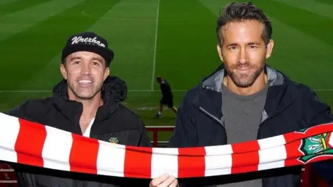 PA Media Rob Mac and Ryan Reynolds when they became owners of Wrexham football club in February 2021, standing in the foreground of the football stadium, holding a red-and-white striped scarf stretched across the lower part of the frame.
